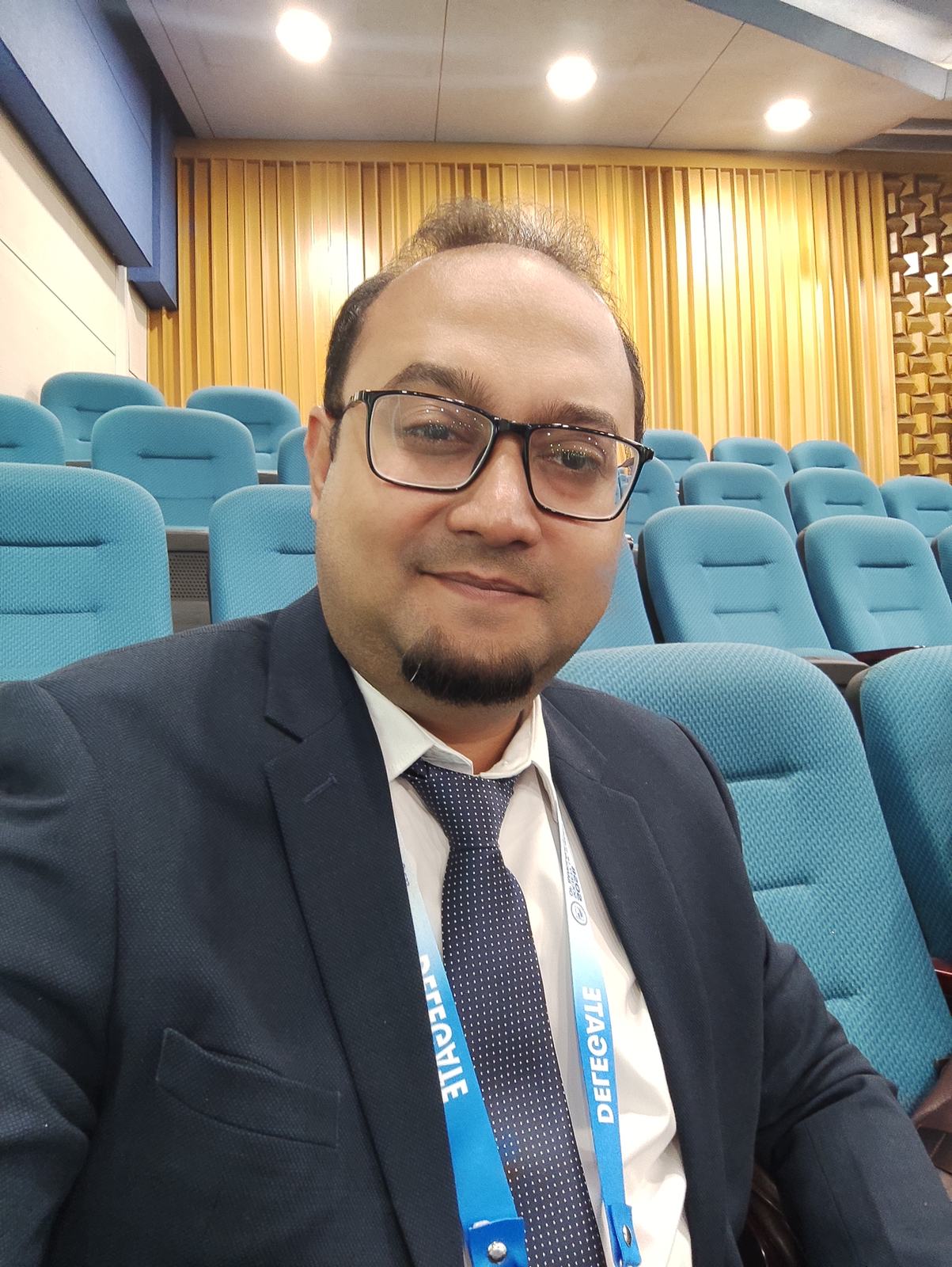 Dr. Murshidul Arefin seated at a medical conference discussing graded operative steps training in an auditorium.